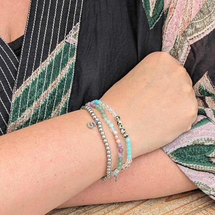 A silver stainless steel PVD beaded stretch bracelet with alphabet letter charm, stacked with other gemstone bracelets.